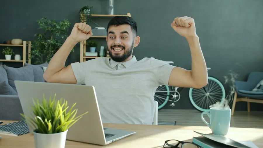 A man at his laptop raising both fists, happy with his day.