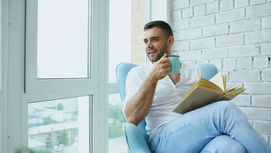 A man smiling by a bright window with coffee and a book, in no hurry.